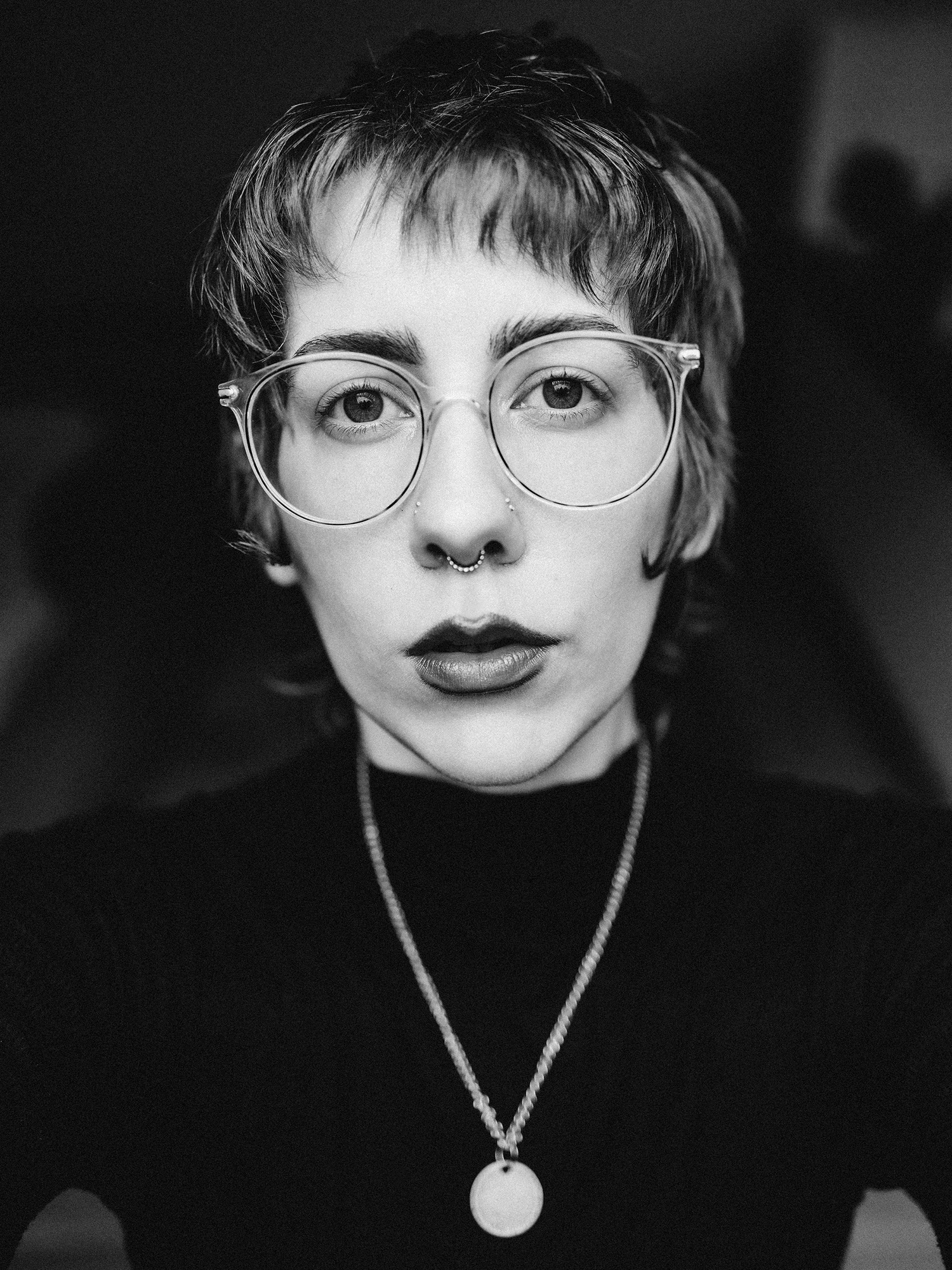 A black and white portrait of Ro Tatchi wearing glasses and a bold lip. They look into the camera pensively and with a serious demeanor.