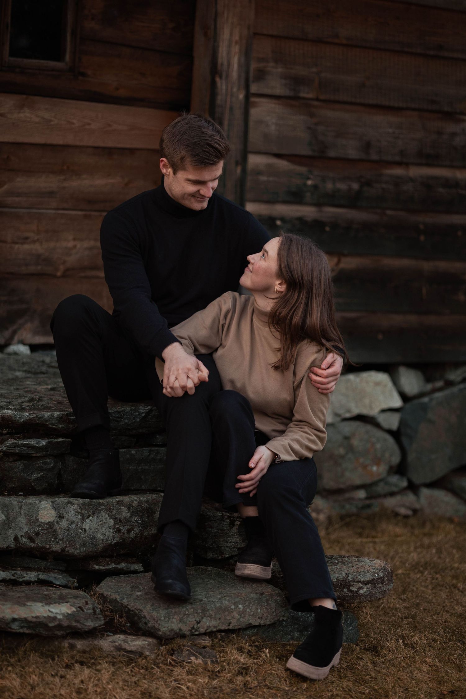 A couple shares a tender moment while sitting on stone steps by a wooden building.