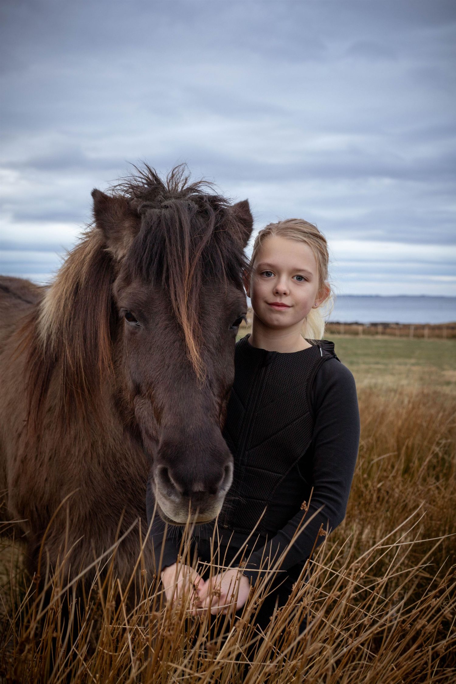 A dark brown Icelandic horse stands close to someone in black clothing against a moody sky.