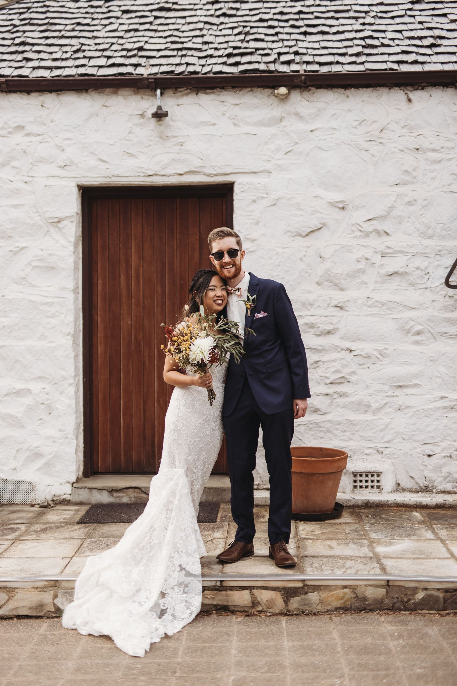 Newlyweds pose in front of white stone wall with wooden door holding wedding bouquet.