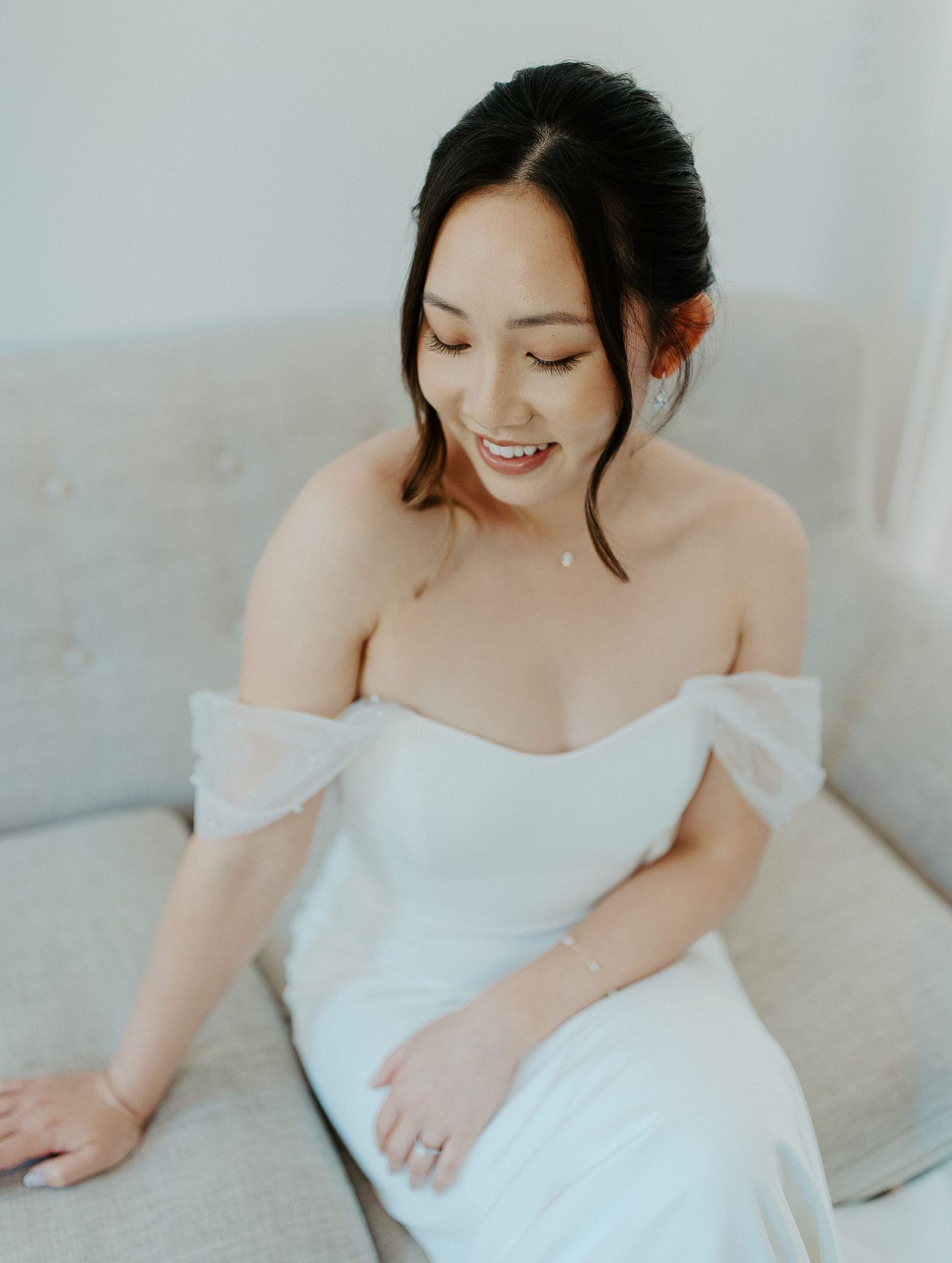 A bride in an off-shoulder white dress smiles sweetly while seated on a couch.