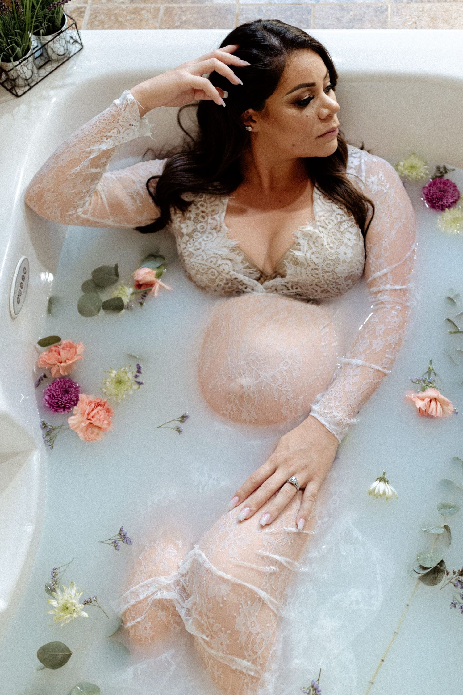 Ethereal maternity milk bath portrait with lace dress and scattered floral petals.
