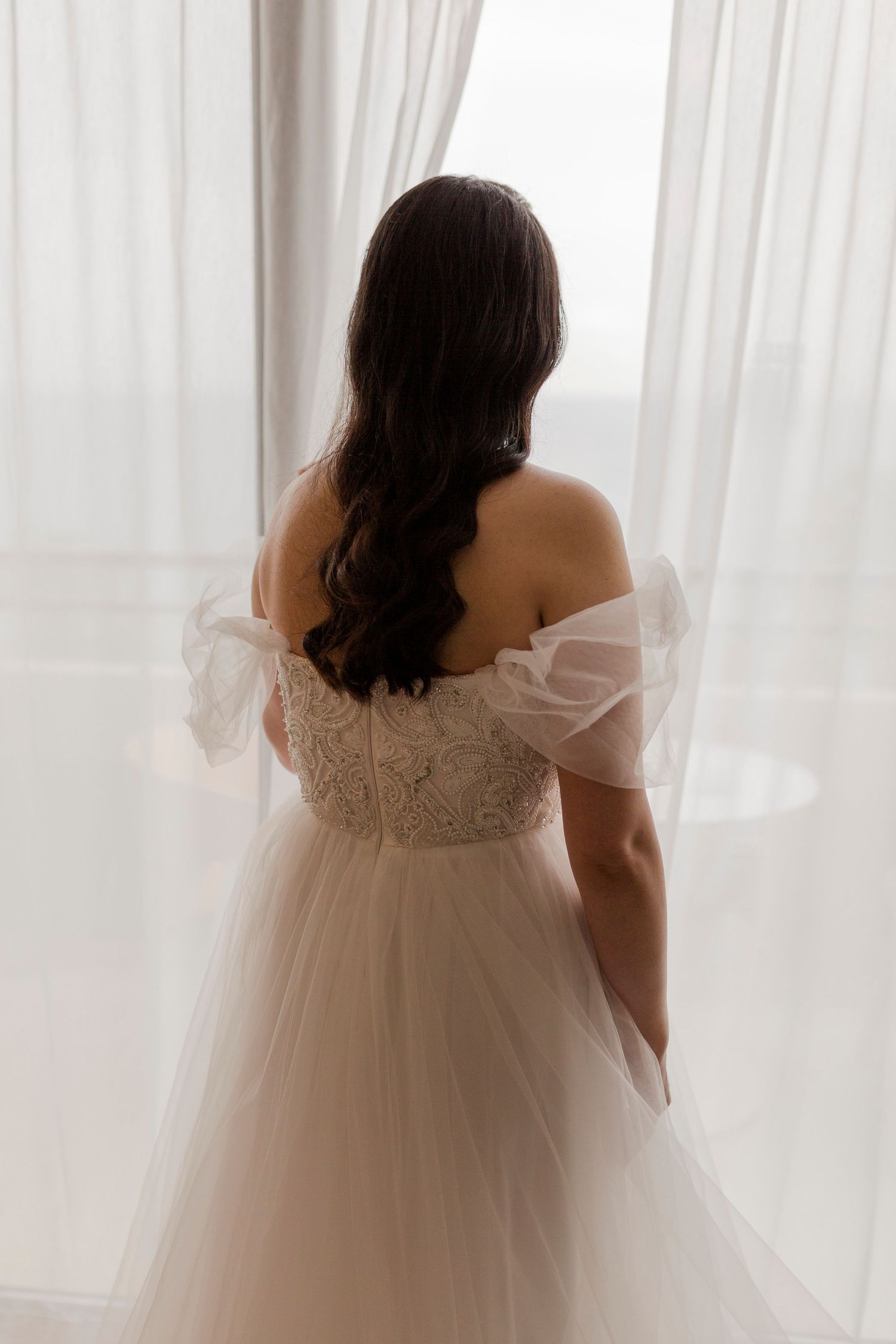 A woman in a wedding dress stands with her back to the camera, looking out sheer curtained windows.